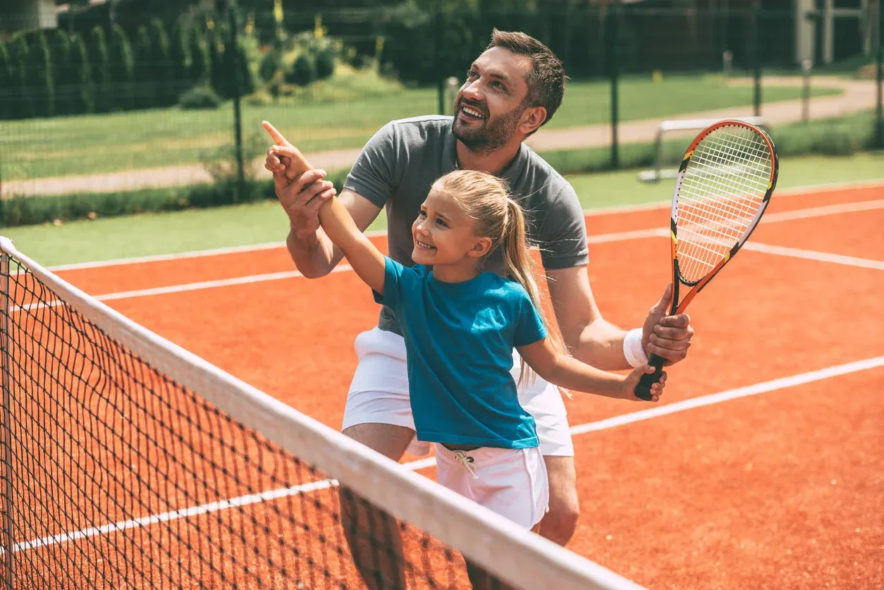 Cheerful father in sports clothing teaching his daughter to play tennis while both standing on tennis court